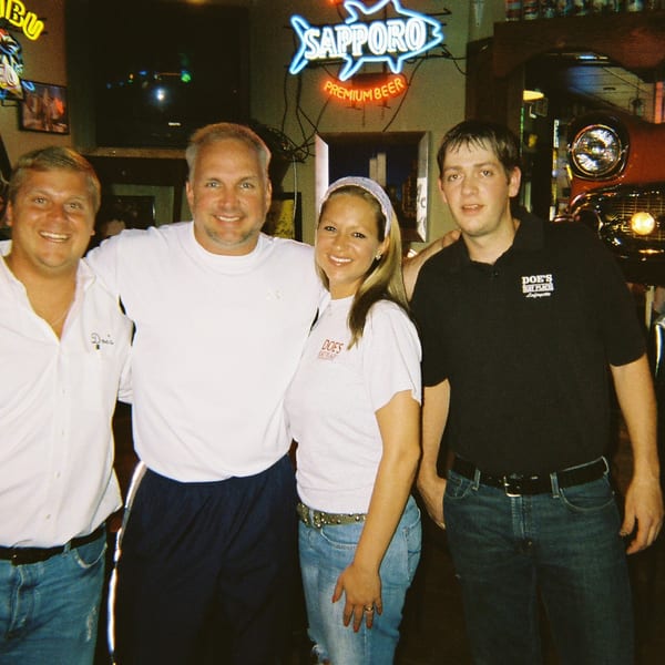 Four smiling people, some wearing 'Doe's Eat Place' shirts, stand in a restaurant with neon signs and a classic car.