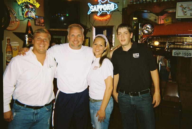 Four smiling people, some wearing 'Doe's Eat Place' shirts, stand in a restaurant with neon signs and a classic car.