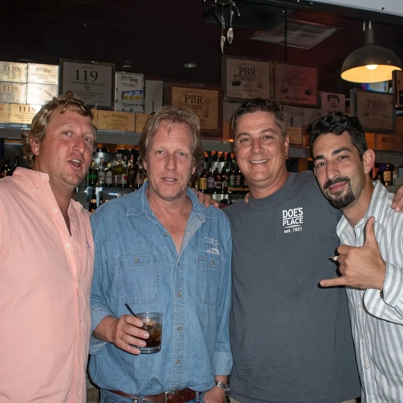 Four men pose for a photo in a bar with bottles and wooden crates in the background.