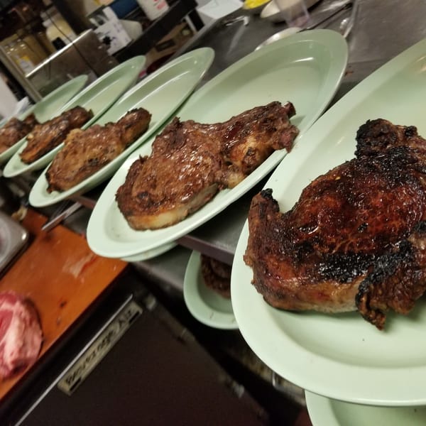 Five perfectly grilled steaks on light green plates lined up in a restaurant kitchen.