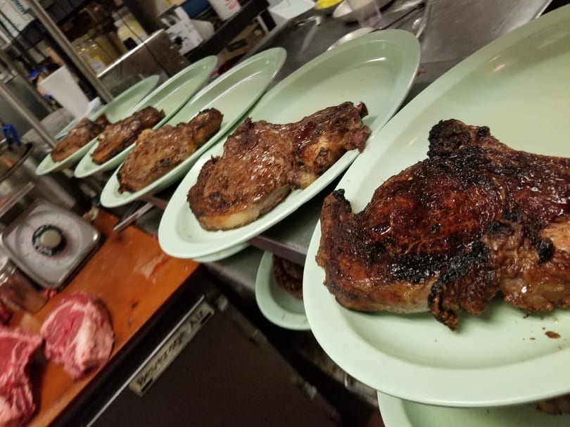 Five perfectly grilled steaks on light green plates lined up in a restaurant kitchen.