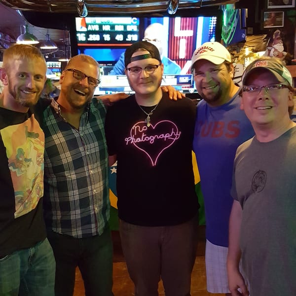 Five smiling men pose together in a lively bar with a classic car front and TV screen.