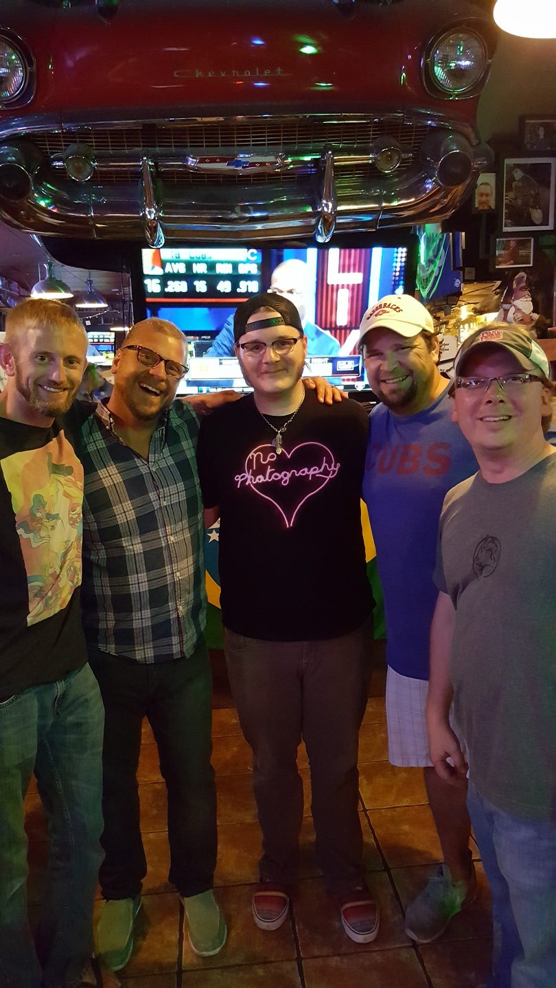 Five smiling men pose together in a lively bar with a classic car front and TV screen.