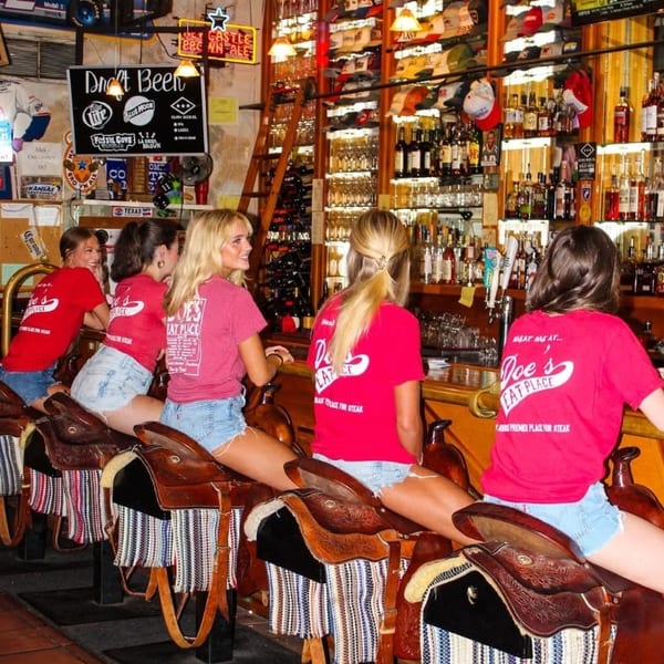 Young women sit on saddle bar stools at a rustic bar, wearing "Doe's Eat Place" shirts.