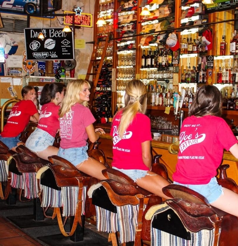 Young women sit on saddle bar stools at a rustic bar, wearing "Doe's Eat Place" shirts.