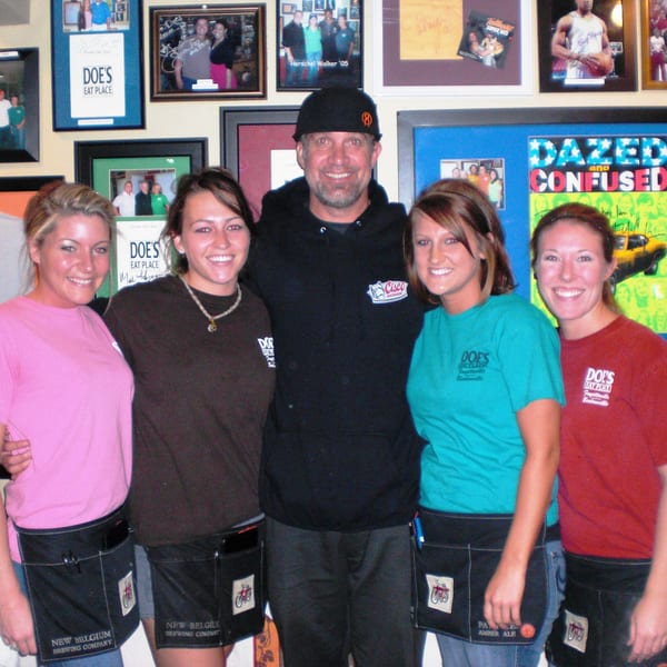 A man and four smiling women, likely servers, stand in front of a memorabilia-filled wall at Doe's Eat Place.