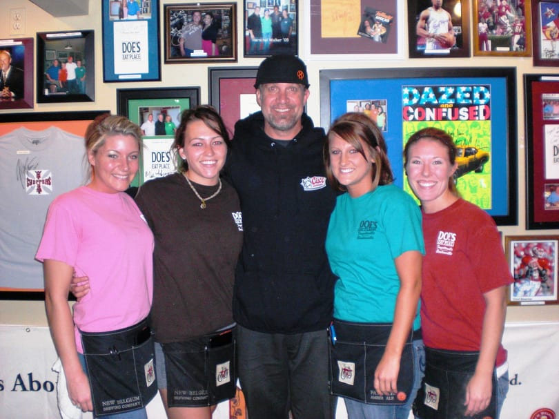 A man and four smiling women, likely servers, stand in front of a memorabilia-filled wall at Doe's Eat Place.
