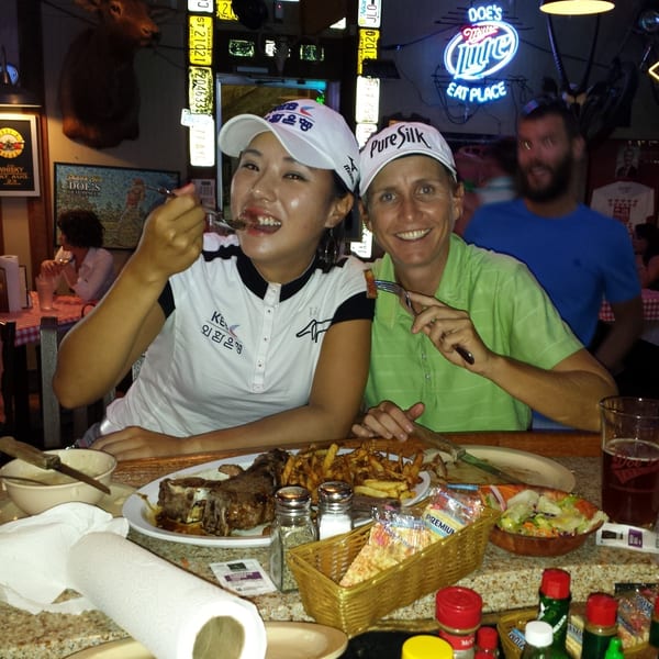 Two women smile while eating steak and fries at a rustic restaurant with a "Doe's Eat Place" neon sign.