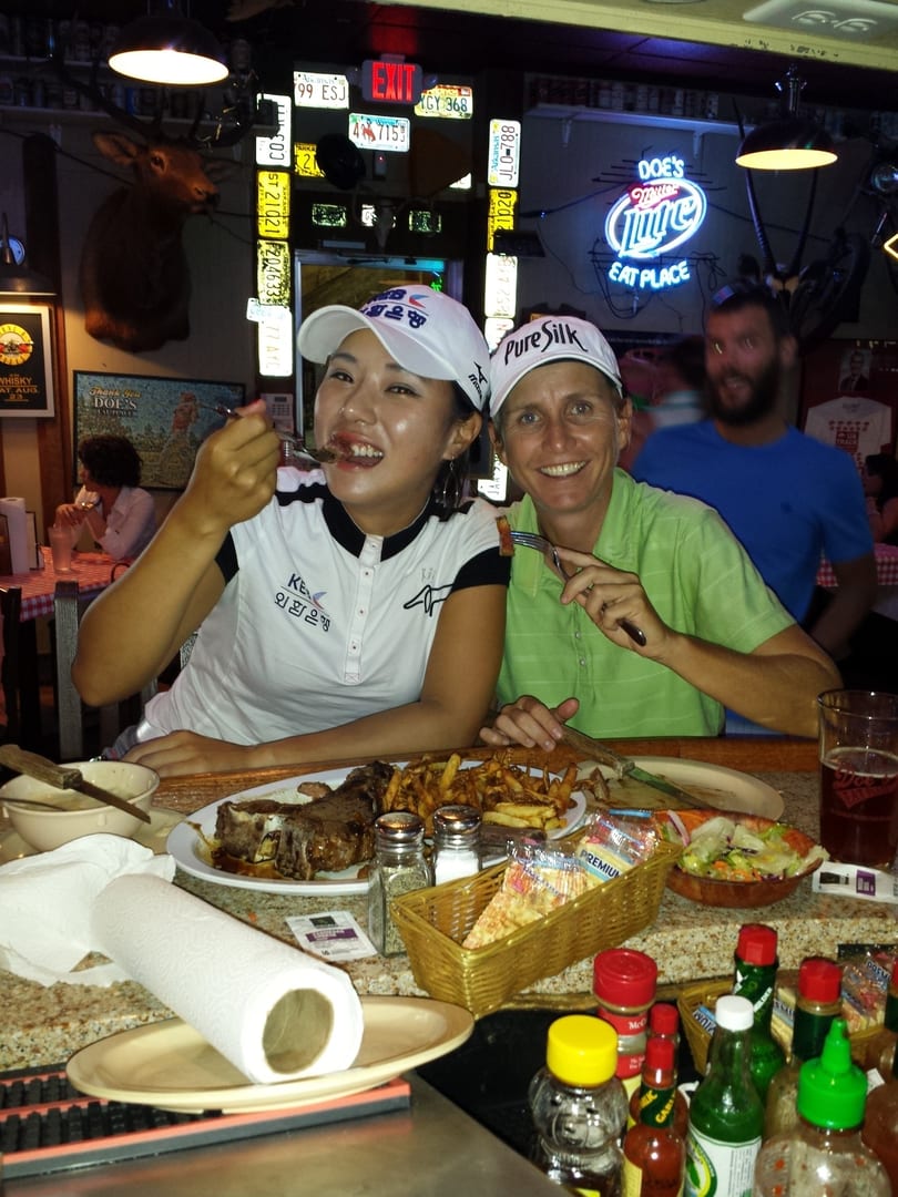 Two women smile while eating steak and fries at a rustic restaurant with a "Doe's Eat Place" neon sign.