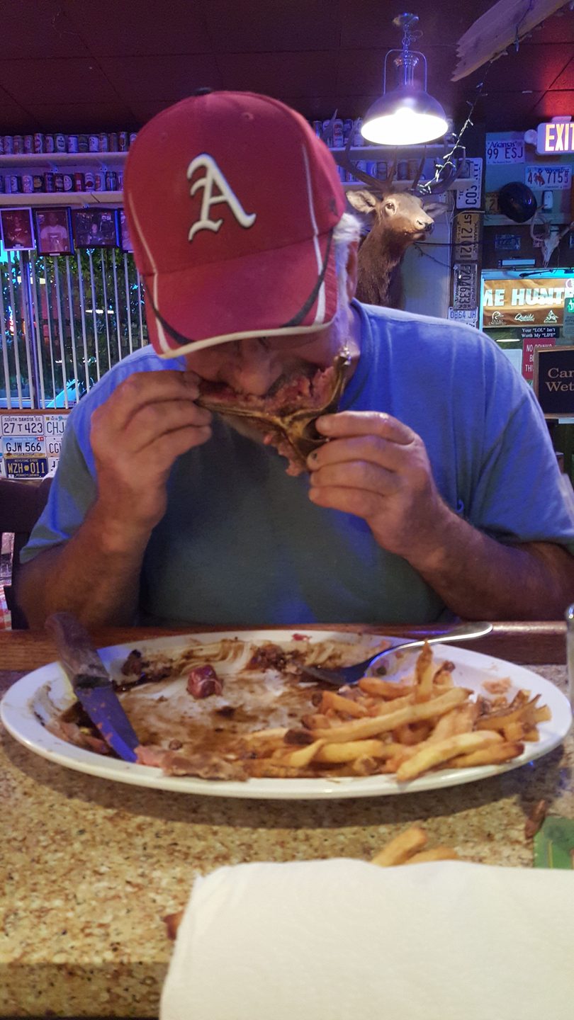 Man in a red baseball cap eating ribs and fries at a casual restaurant with a deer head mount.