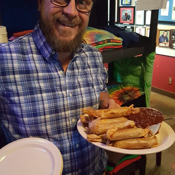 Smiling man in plaid shirt holding a plate of tamales and sauce at Doe's Eat Place.