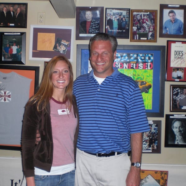A smiling man and woman stand in front of a wall covered with framed sports and celebrity memorabilia.