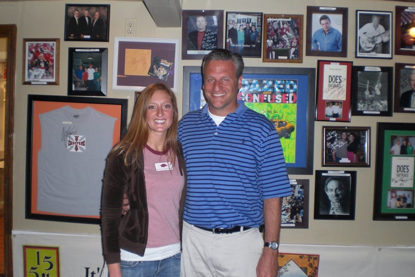 A smiling man and woman stand in front of a wall covered with framed sports and celebrity memorabilia.