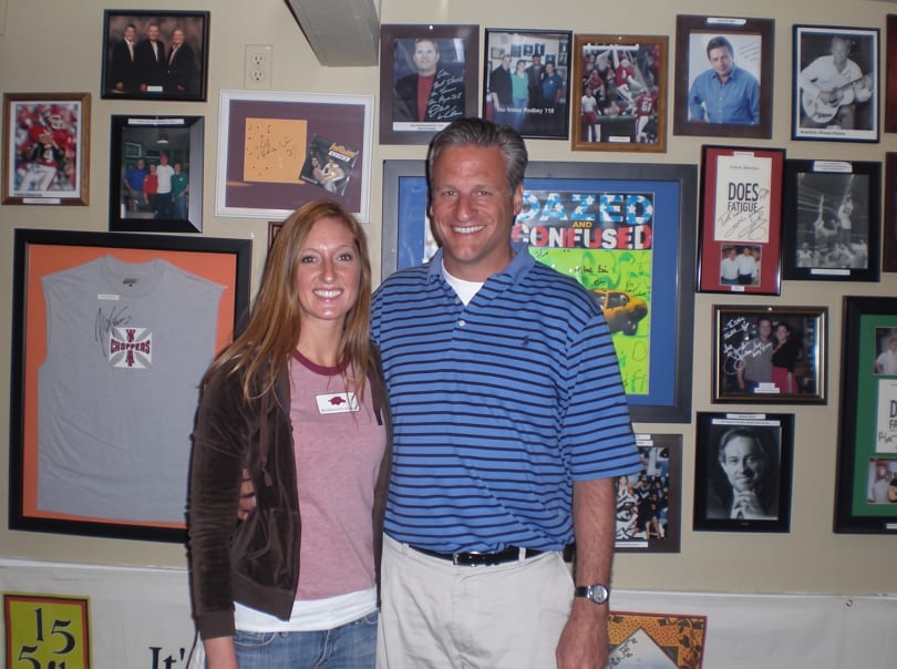 A smiling man and woman stand in front of a wall covered with framed photos and memorabilia.