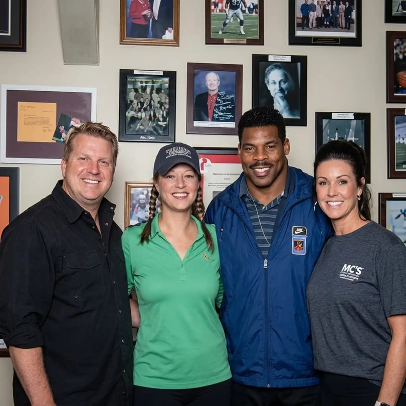 Four smiling people, including Herschel Walker, pose for a photo in front of a wall of framed pictures.