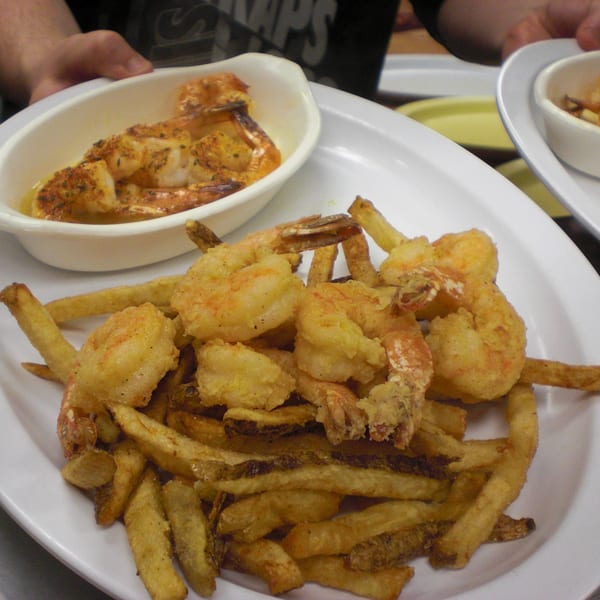 A white plate holds fried shrimp and french fries, with a small dish of seasoned shrimp in the background.