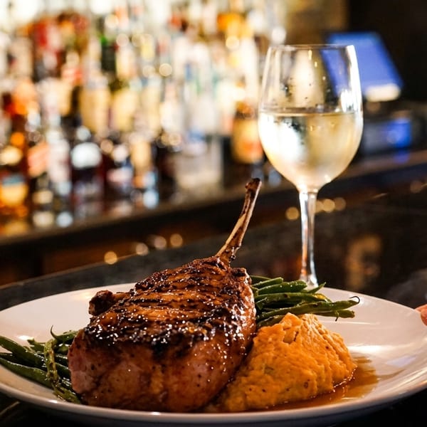 A hand placing a plate with a grilled pork chop, green beans, and mashed sweet potatoes on a bar.
