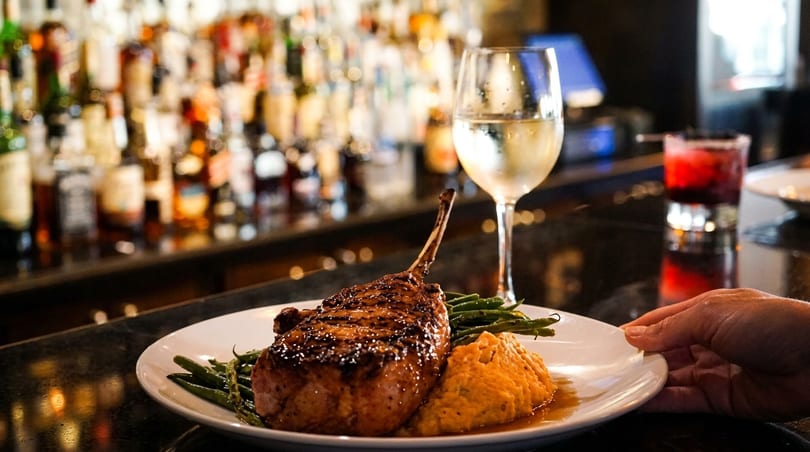 A hand placing a plate with a grilled pork chop, green beans, and mashed sweet potatoes on a bar.