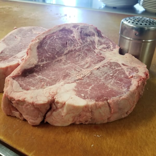 Two thick raw steaks on a wooden cutting board with a salt shaker.