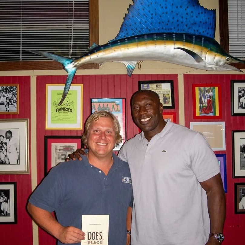 Two men smiling, one holding a "Doe's Place" menu, in a restaurant with a large fish decoration.