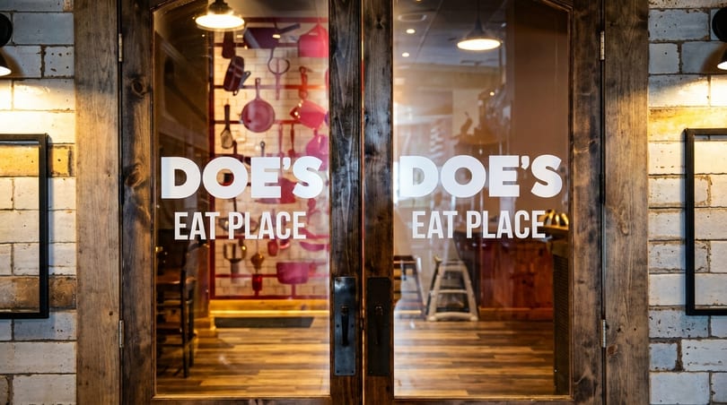 The entrance to Doe's Eat Place, with the restaurant name on glass doors and a brick wall.