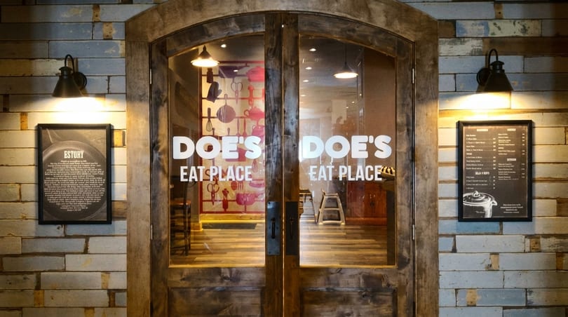 The entrance to Doe's Eat Place, featuring wooden doors, a rustic brick wall, and menu boards.