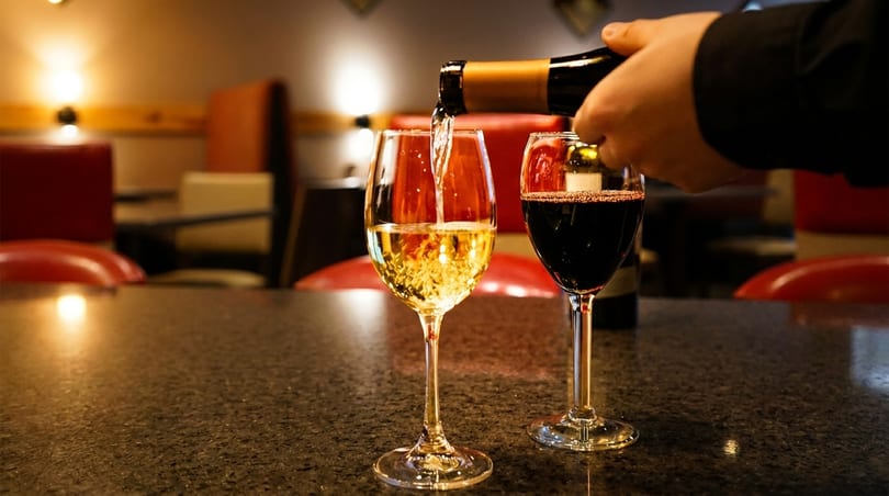 A hand pours white wine into a glass next to a full glass of red wine on a dark table.