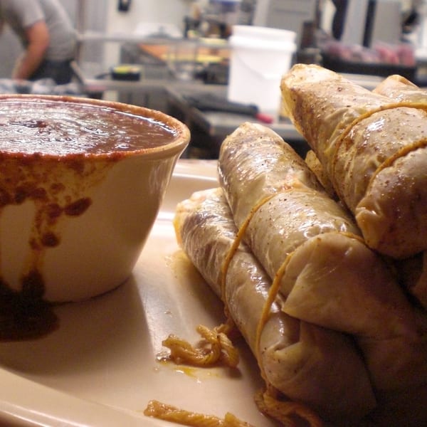 A plate of tamales and a cup of chili with a person working in the background.