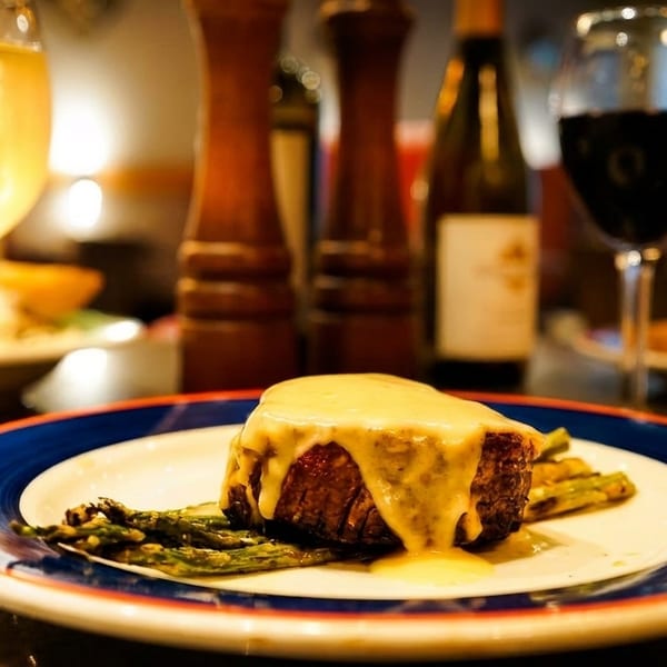 Steak with sauce and asparagus on a plate, with wine glasses and bottles in the background.