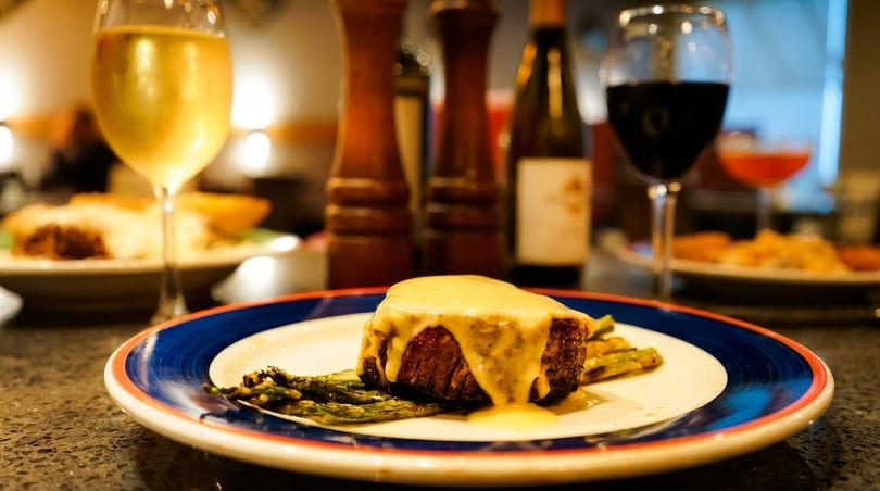 Steak with sauce and asparagus on a plate, with wine glasses and bottles in the background.