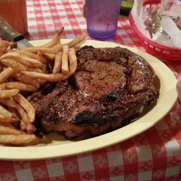 Large grilled steak and french fries on a yellow plate, with a hand giving a thumbs-up on a checkered table.
