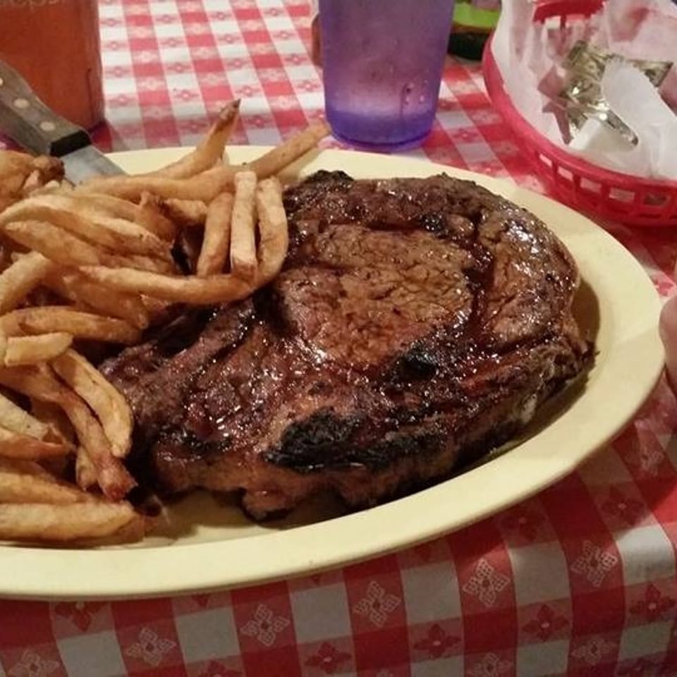 Large grilled steak and french fries on a yellow plate, with a hand giving a thumbs-up on a checkered table.