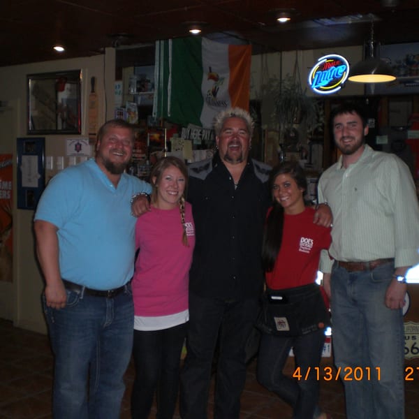 Guy Fieri poses with four smiling people in a dimly lit bar with an Irish flag.