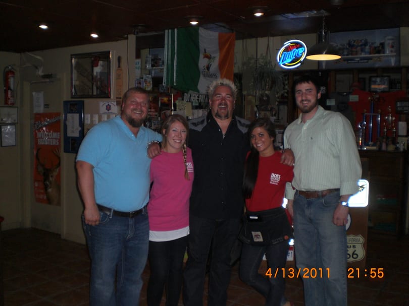 Guy Fieri poses with four smiling people in a dimly lit bar with an Irish flag.