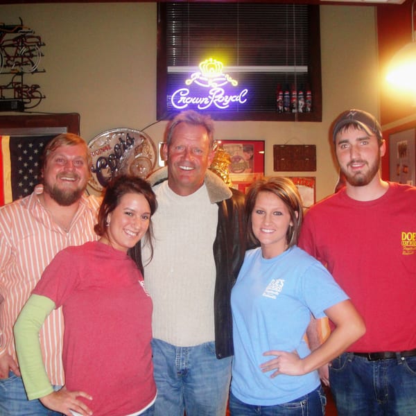 Five smiling people, some wearing "Doe's Eat Place" shirts, in a bar with neon signs.
