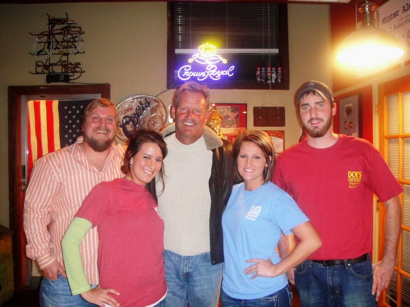 Five smiling people, some wearing "Doe's Eat Place" shirts, in a bar with neon signs.