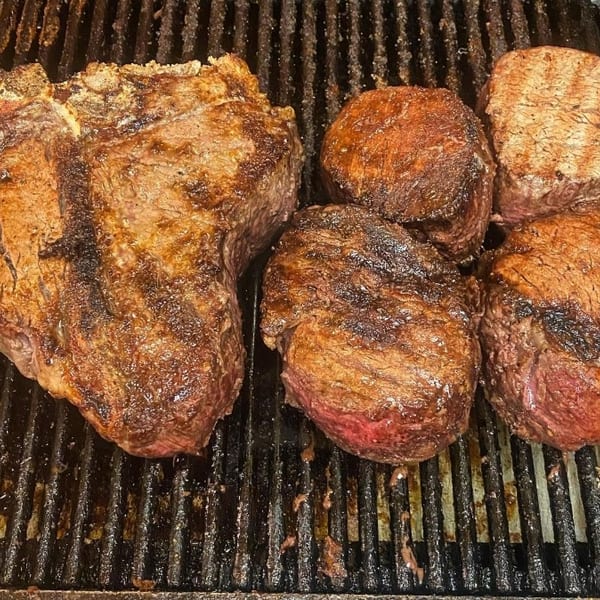 Sizzling T-bone steak and four smaller cuts cooking on a dark, well-used grill.