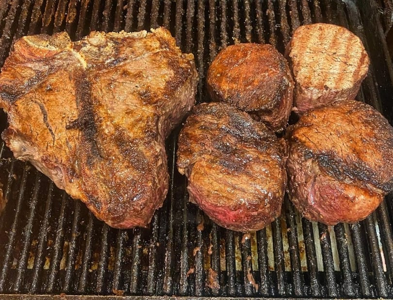 Sizzling T-bone steak and four smaller cuts cooking on a dark, well-used grill.