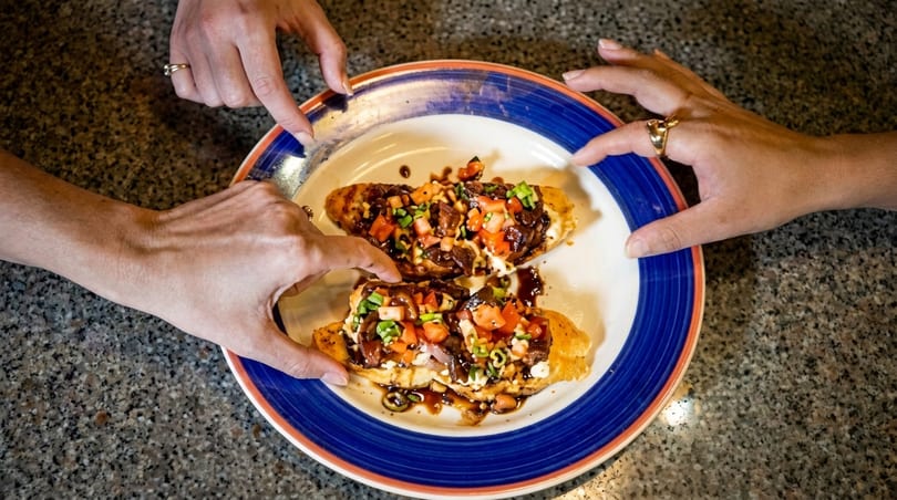 Four hands reaching for two bruschetta pieces topped with meat, tomatoes, and green onions.