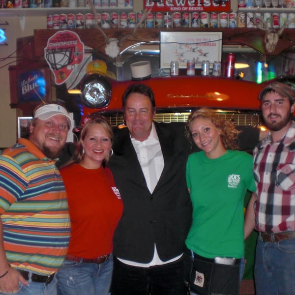 Five smiling people, including a man in a suit, pose at Doe's Eat Place with bar decor.
