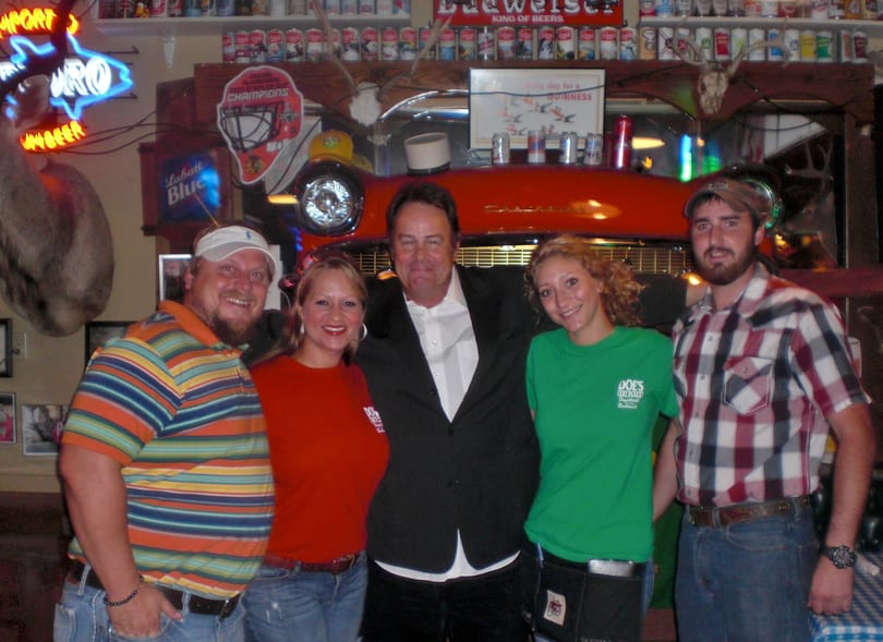 Five smiling people, including a man in a suit, pose at Doe's Eat Place with bar decor.
