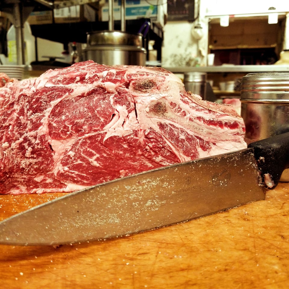 Raw, seasoned beef roast on a wooden cutting board with a large knife in a restaurant kitchen.