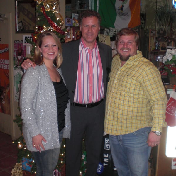 Three smiling people, including Will Ferrell, pose in a festive bar with a Christmas tree and Irish flag.