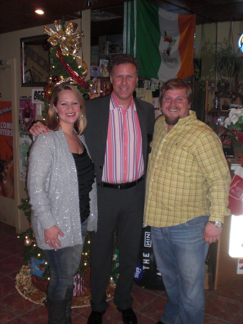 Three smiling people, including Will Ferrell, pose in a festive bar with a Christmas tree and Irish flag.