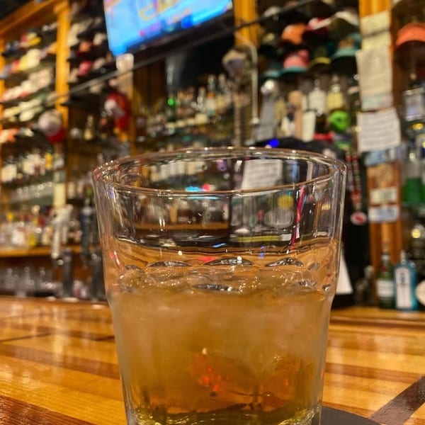 Glass of amber drink with ice on a wooden bar counter, with a TV and bottles in the background.