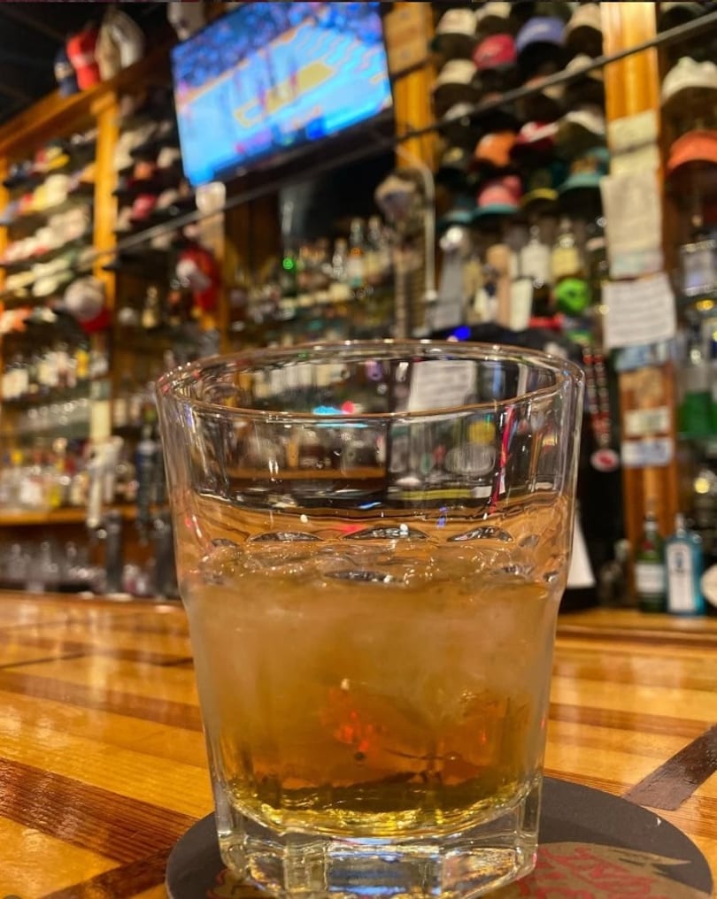 Glass of amber drink with ice on a wooden bar counter, with a TV and bottles in the background.