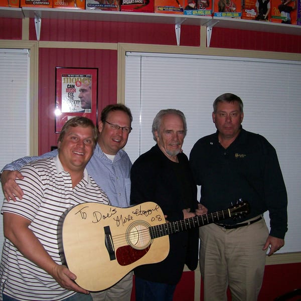 Four men pose with a signed acoustic guitar by Merle Haggard in a room with sports memorabilia.
