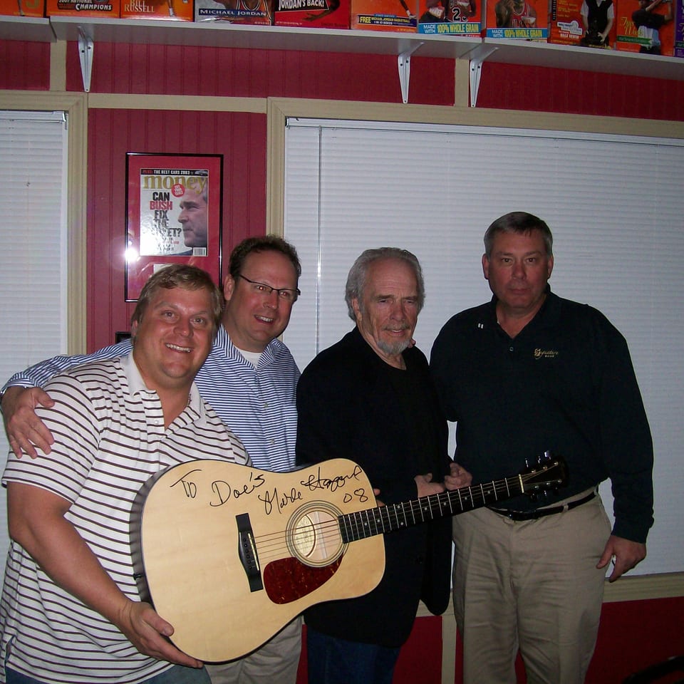 Four men pose with a signed acoustic guitar by Merle Haggard in a room with sports memorabilia.
