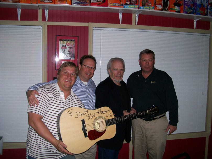 Four men pose with a signed acoustic guitar by Merle Haggard in a room with sports memorabilia.