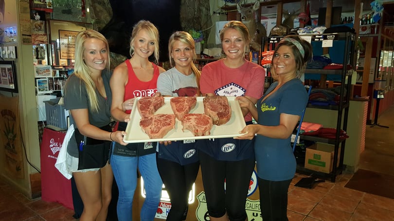Five smiling women, likely waitstaff at Doe's Eat Place, proudly display a large tray of raw steaks.